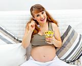 Smiling beautiful pregnant woman sitting on sofa with fruit salad  in hand
