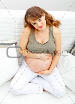 Smiling beautiful pregnant female sitting on sofa and touching her belly
