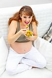 Smiling beautiful pregnant woman sitting on couch and eating fruit salad
