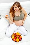 Smiling  beautiful pregnant woman sitting  on sofa and  holding fruit in hand

