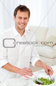 Joyful man serving salad standing at a table in the living room