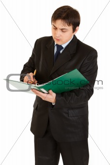 Serious young businessman holding folder in hand and making notes in document
