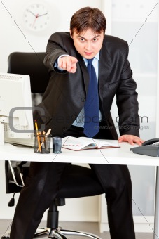 Angry  modern businessman standing at office desk and pointing finger on you
