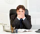 Modern businessman sitting at office desk and making speak no evil gesture

