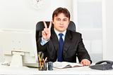 Confident modern businessman sitting at office desk and showing victory gesture
