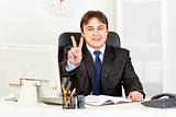 Smiling modern businessman sitting at office desk and showing victory gesture
