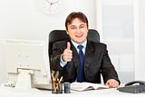 Smiling modern businessman sitting at office desk and showing thumbs up gesture
