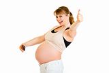 Laughing pregnant woman photographing her belly and showing thumbs up gesture
