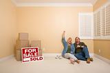 Excited Couple Relaxing on Floor Near Boxes and Sold Real Estate Signs in Empty Room.