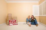 Excited Couple With New Keys Relaxing on Floor Near Boxes and Sold Real Estate Signs in Empty Room.