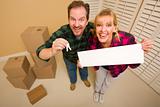 Goofy Couple Holding Keys and Blank Sign in Room with Packed Cardboard Boxes.