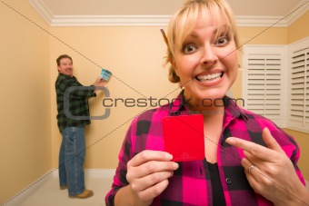 Fun Happy Couple Comparing Paint Colors in Empty Room - Woman Large, in Front, Man Smaller, Behind.