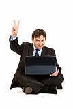 Smiling  businessman sitting on floor with laptops and showing victory gesture
