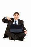 Smiling businessman sitting on floor and pointing finger at laptop
