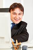 Smiling modern businessman sitting at office desk and looking from computer monitor

