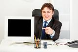 Smiling  businessman sitting at office desk and pointing finger at  monitor with blank screen
