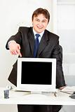 Smiling  businessman standing behind office desk and pointing finger at  monitor with blank screen
