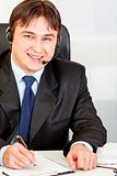 Smiling  businessman with  headset sitting at office desk and taking notes on paper
