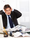  Shocked young businessman sitting at office desk  being overloaded with loads of work 

