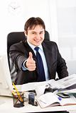 Pleased businessman sitting at office desk and showing  thumbs up gesture
