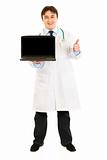 Smiling  doctor holding laptops with blank screen and showing  thumbs up gesture
