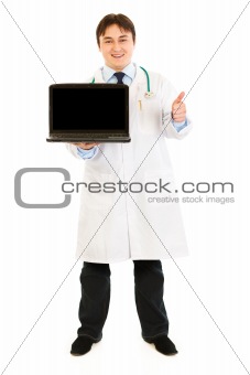 Smiling  doctor holding laptops with blank screen and showing  thumbs up gesture
