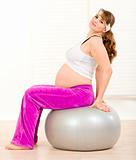 Smiling beautiful pregnant woman doing pilates exercises on gray ball at home
