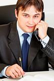 Smiling modern businessman sitting at office desk and talking on phone
