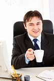 Smiling businessman  sitting at office desk  and holding cup of tea in hand
