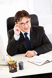 Smiling  businessman sitting at office desk and straightening eyeglasses

