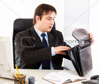 Shocked businessman sitting at office desk and searching something in briefcase 
