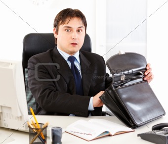 Shocked modern businessman sitting at office desk and holding open suitcase in hands
