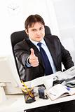 Successful businessman sitting at office desk and showing thumbs up gesture
