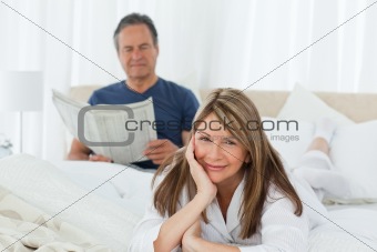 Smiling woman looking at the camera  while her husband is readin