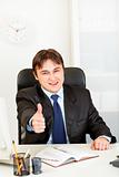 Pleased modern businessman sitting at office desk and showing thumbs up gesture
