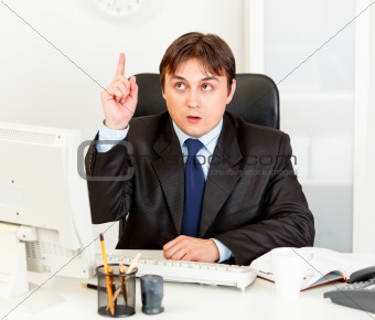 Concentrated businessman with rised finger sitting at office desk. Idea gesture
