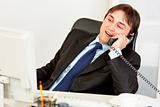 Pleased modern businessman sitting at office desk and talking on phone
