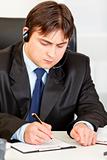 Concentrated  businessman with  headset sitting at office desk and taking notes on paper
