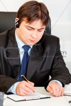 Concentrated  businessman with  headset sitting at office desk and taking notes on paper
