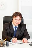 Smiling  businessman with  headset sitting at office desk and taking notes on paper
