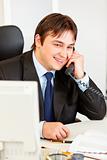 Smiling modern businessman sitting at office desk and talking on phone
