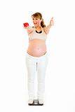 Pleased pregnant woman standing on weight scale and showing thumbs up
