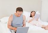 Manworking on his laptop while his wife is reading a book on the
