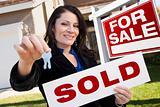 Happy Attractive Hispanic Woman Holding Sold Real Estate Sign and Keys in Front For Sale Real Estate Sign and House.
