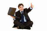 Smiling  businessman sitting on floor with laptop showing thumbs up gesture
