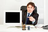 Authoritative businessman sitting at office desk and pointing finger at  monitor with blank screen
