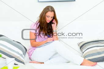 Concentrated pretty woman talking on phone and making notes in pad
