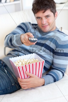 Relaxed young man eating popcorn and holding a remote lying on t