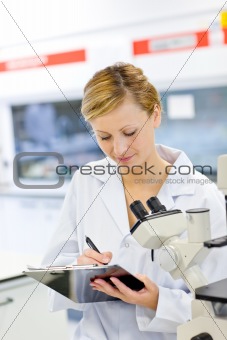 Serious female scientist writing on her clipboard in front of a 