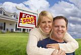 Happy Couple Hugging in Front Yard with Sold Real Estate Sign and House.